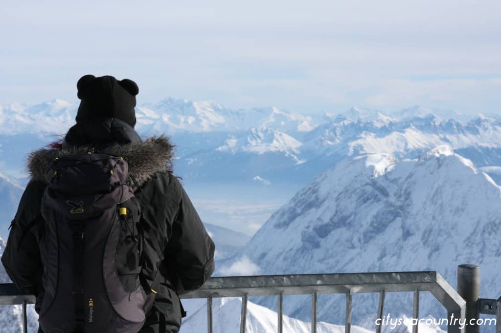 a woman with a black backpack, jacket and black hat with bear ears looking down the Zugspitze mountain, Germany' highest mountain covered in snow