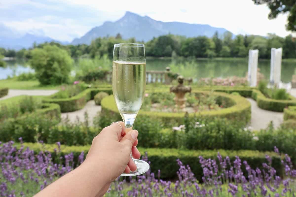 A hand holding a champagne glass at the castle Schloss Leopoldskron terrace with a view of the garden, the pond, The Sound of Music filming location view, and the impressive Untersberg mountain in the background.