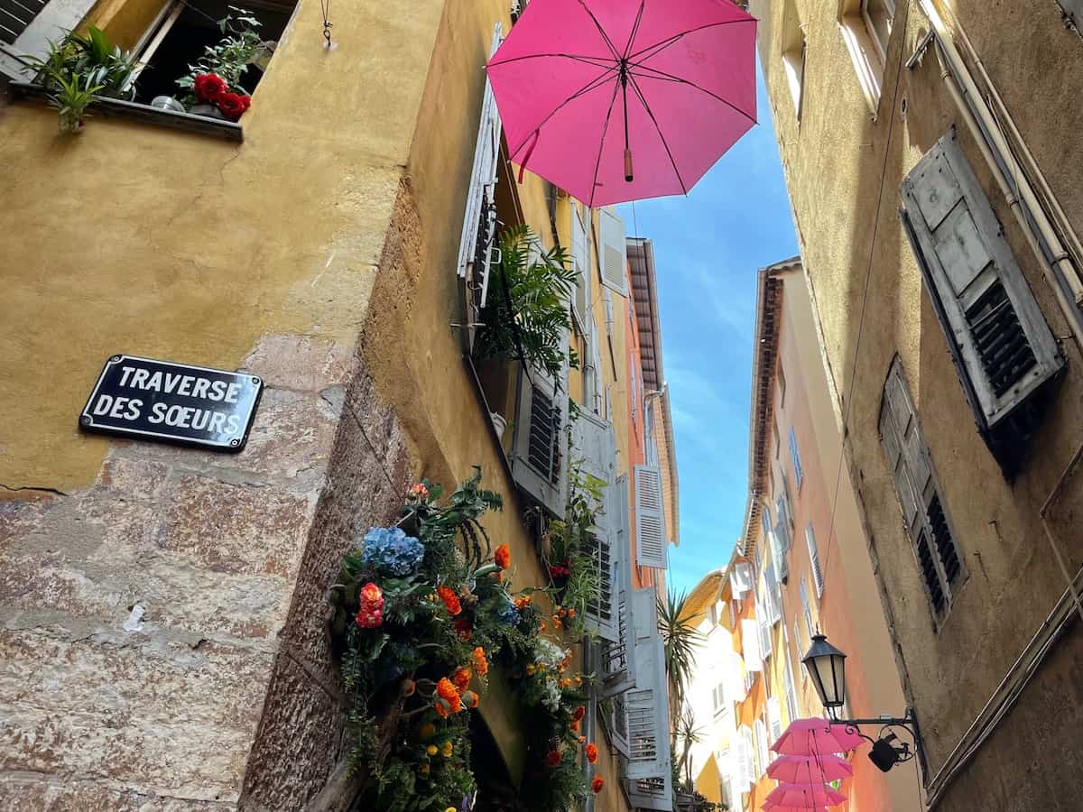 Bild einer engen Gasse in Grasse, die mit einem rosa Regenschirm und bunten Kunstblumen geschmückt ist während eines Tagesausflug von Nizza aus