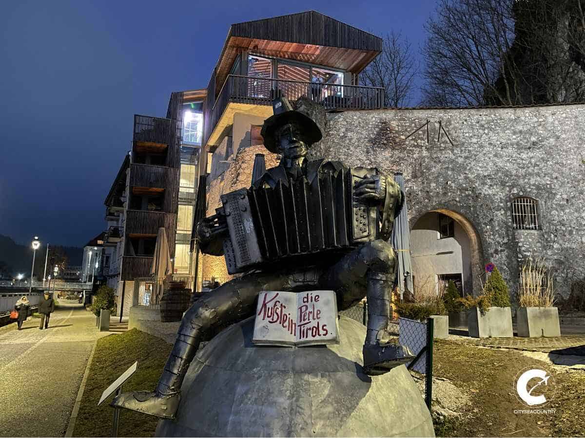Denkmal des Karl Ganzer auf einer Kugel sitzend mit Ziehharmonika und Tiroler Hut vor Stadtmauer, Auracher Löchl bei Nacht
