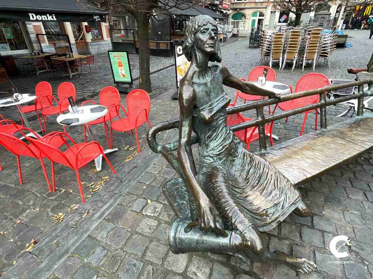 Statue einer sitzenden Kotmadam aus Bronze auf einer Parkbank. In der rechten Hand hält sie eine Kanne.