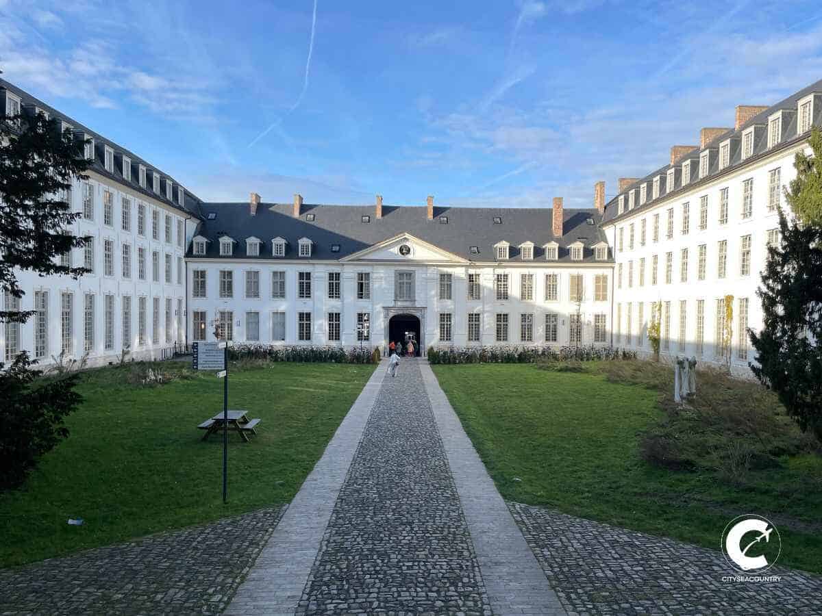 Blick auf den Innenhof und die pompösen weißen Gebäude mit grauem Dach. Ein Weg führt zwischen Grünflächen auf ein großes Tor inmitten des Gebäudes.