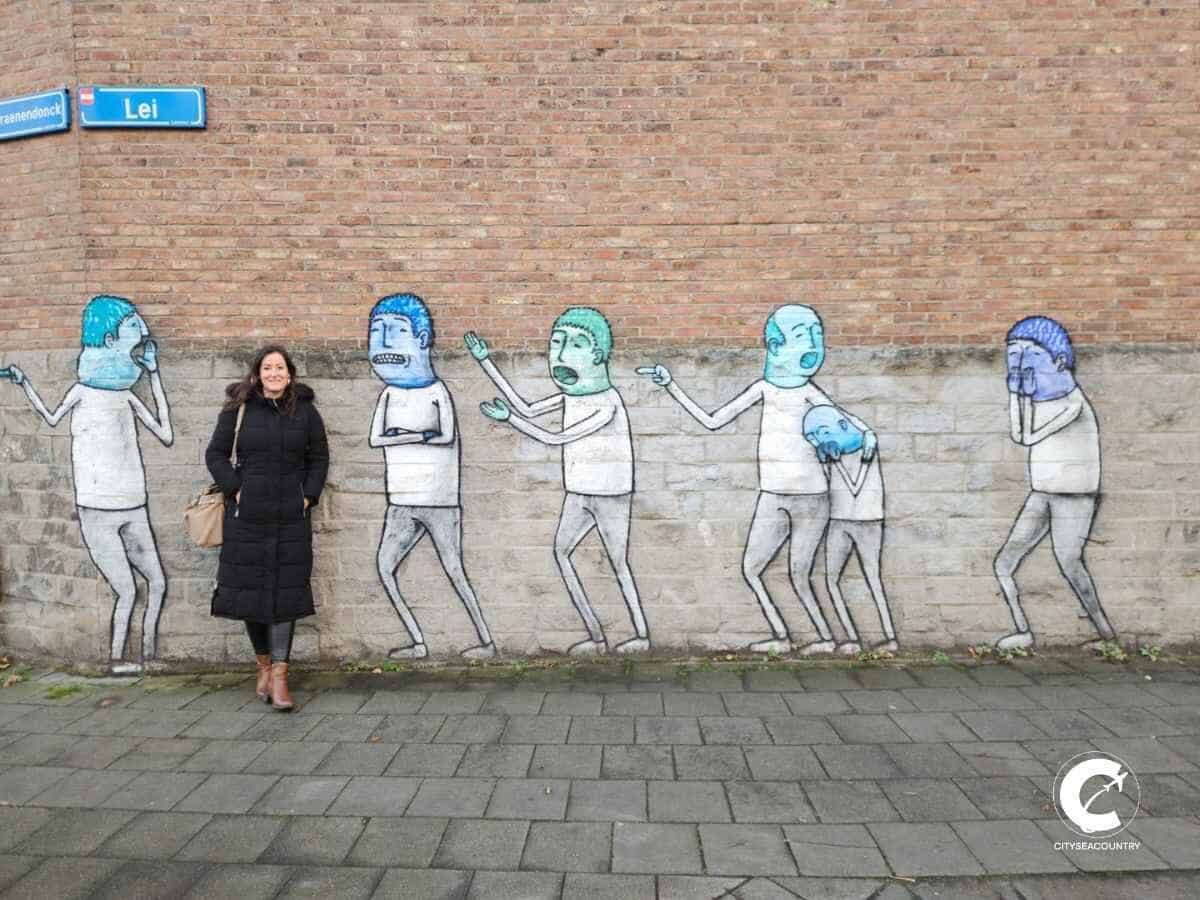 Autorin Christina vor einem Mural mit verschiedenen Personen in weiß und blauen oder türkisfarbenen Haaren vor typischen Haus aus roten Sandsteinen