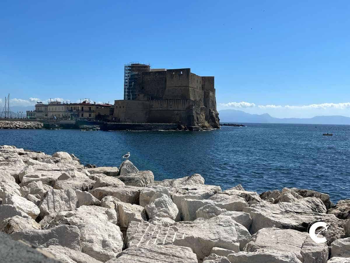 große Felsblöcke an der Uferpromenade mit dem dunkelblauen Meer und der dunkelbraunen Festund Castel Dell'Ovo umgeben vom Meer.