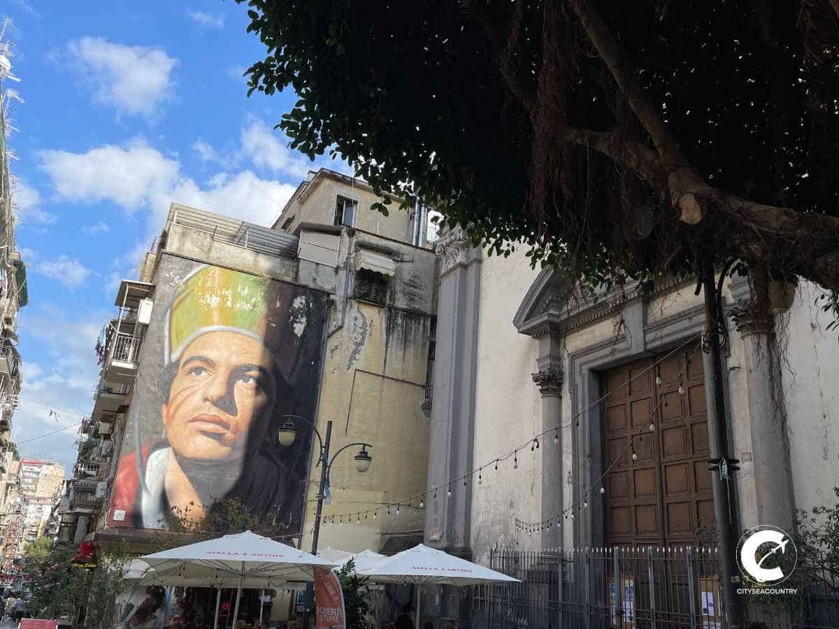 Blick auf eine der vielen Kirchen in der Altstadt von Neapel neben einem Gebäude mit einem großen Mural mit einem realitischen Mann mit bunter Kopfbedeckung