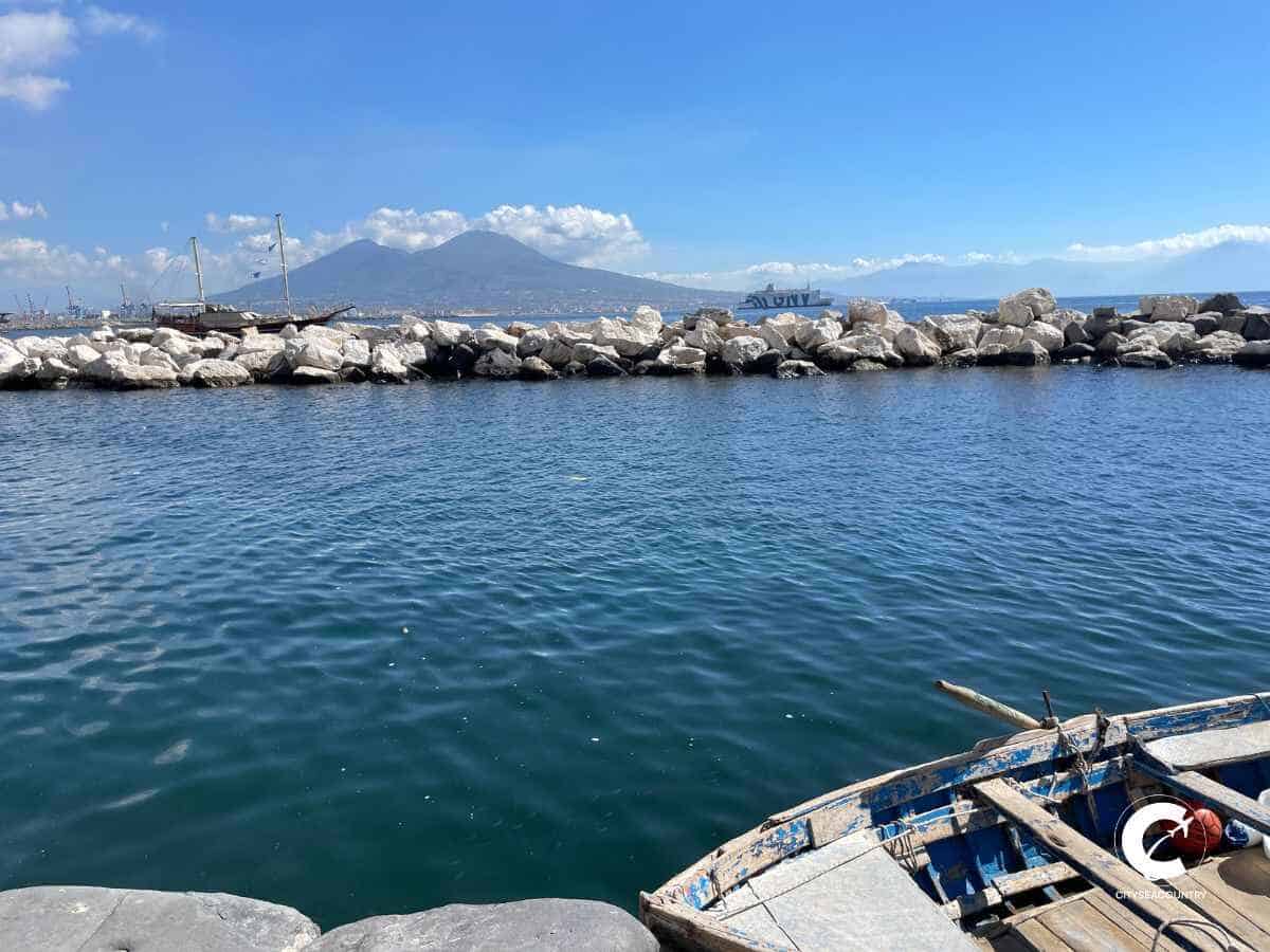 Blick von der Uferpromenade mit einem alten Holzboot an der Kaimauer, dem dunkelblauen Meer und Vesuv im Hintergrund