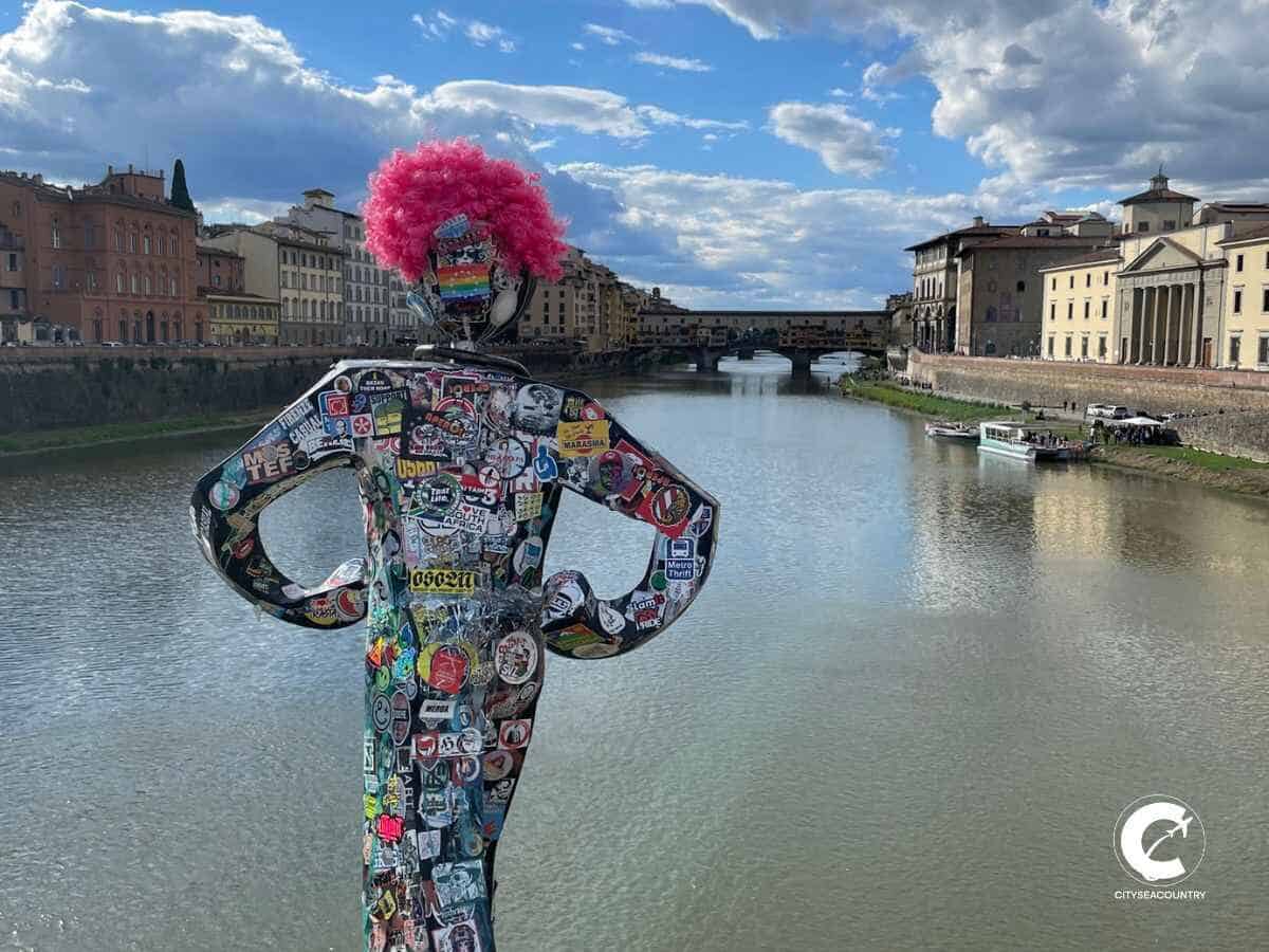 Street Art Skulptur mit Stickern beklebt und einer pinken Perücke oberhalb des Fluss Arno mit Blick auf die Ponte Vecchio.