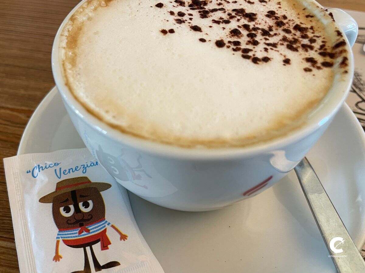 Tasse Cappuccino mit einem Säckchen Zucker auf welchem eine Kaffeebohne als venedischer Gondoliere gezeichnet ist.