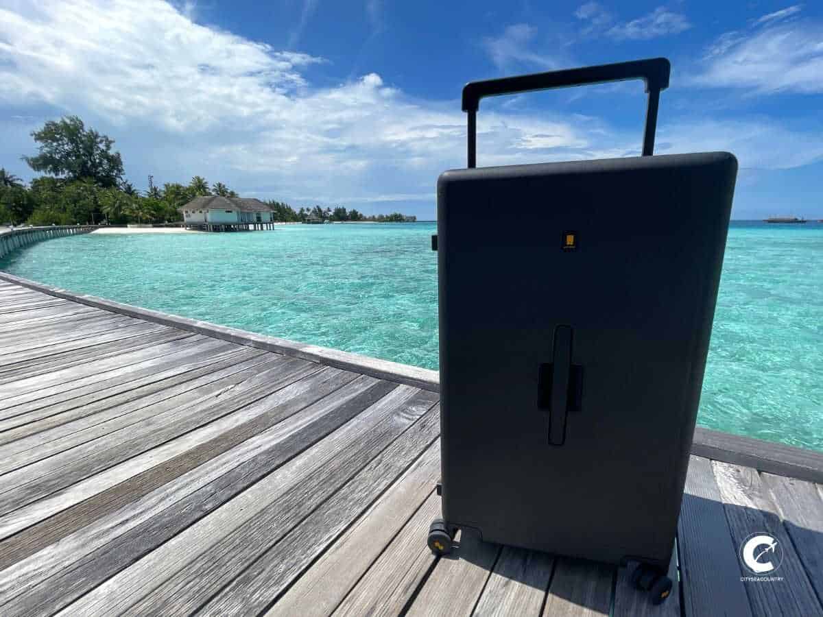 The Level8 Check-In 28'' in black on a wooden pier leading to the water villas in the Maldives. In the background is the turquoise ocean with a view of the island covered wit palm trees.