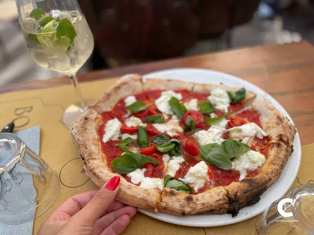 Autorin Christina zieht ein Stück einer Büffelmozzarellapizza mit frischem Basilikum vom Teller. Links hinten ist auf dem Holztisch ein Aperitiv zu sehen.