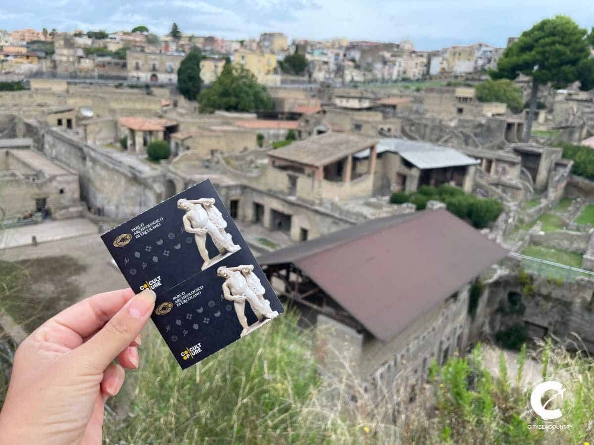 Frau hält Tickets für Herculaneum in der Hand. Im Hintergrund die Ausgrabungsstätte Herculaneum.