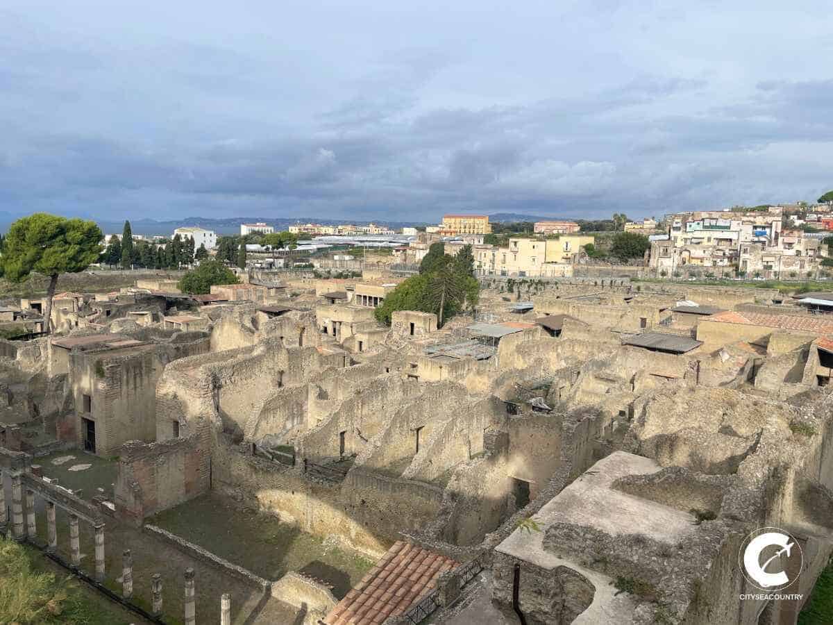 Von der Sonne beleuchtete Ausgrabungsstätte Herculaneum, mit Ruinen von Häusern, einem Garten im Vordergrund. Der Ausblick befindet sich vom oberen Zugangsareal aus.