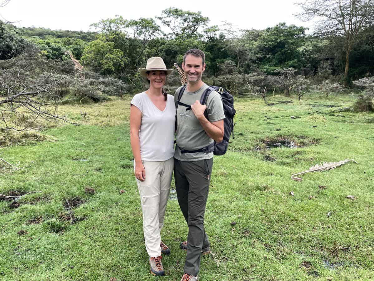 View of Arusha National Park with two giraffes and lush greenery in the background, with author Christina and her partner in the front in safari clothes during a walking safari.